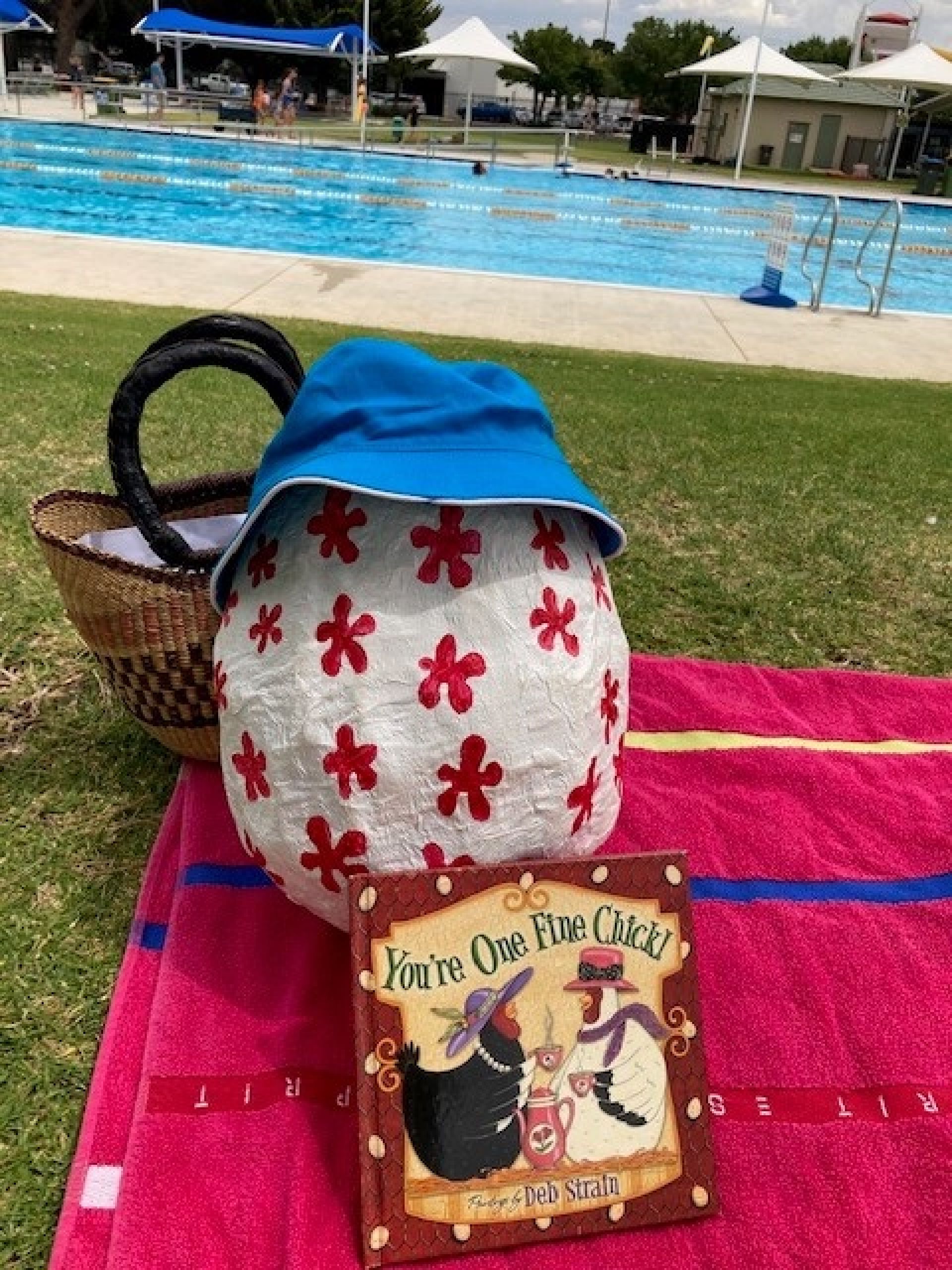 The big egg sitting beside the pool, wearing a hat with a book and a picnic basket.