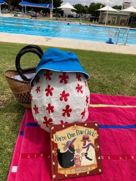 The big egg sitting beside the pool, wearing a hat with a book and a picnic basket.