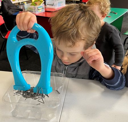 Little boy using a large blue magnet to pick up a collection of small metal objects
