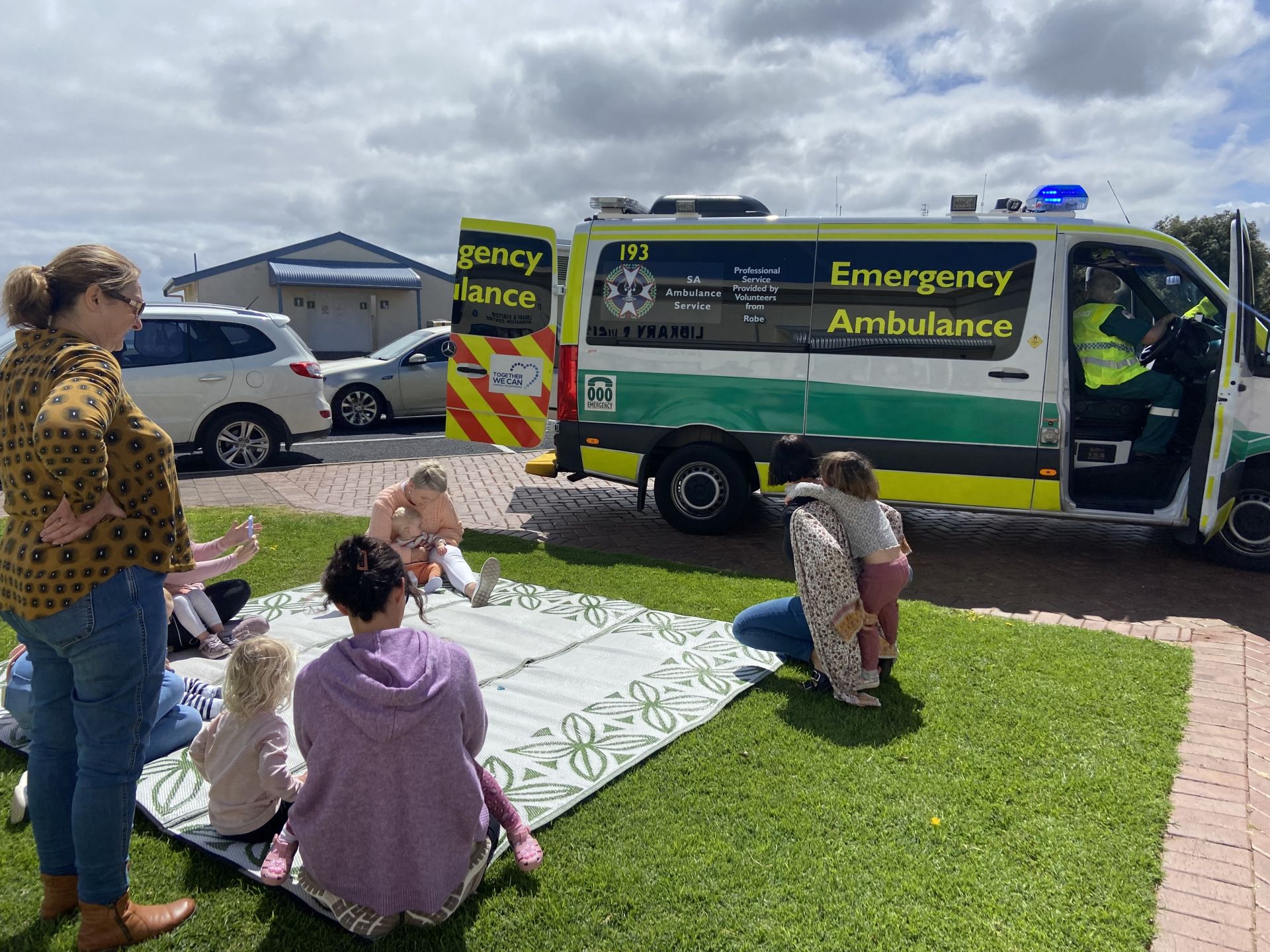 People outside sitting on the grass watching a demonstration by SA Ambulance